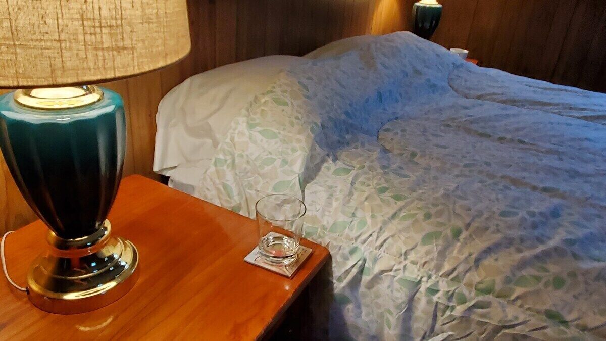 Una cama bien hecha con una colcha estampada, una lámpara de noche y un vaso de agua sobre una mesita de madera.