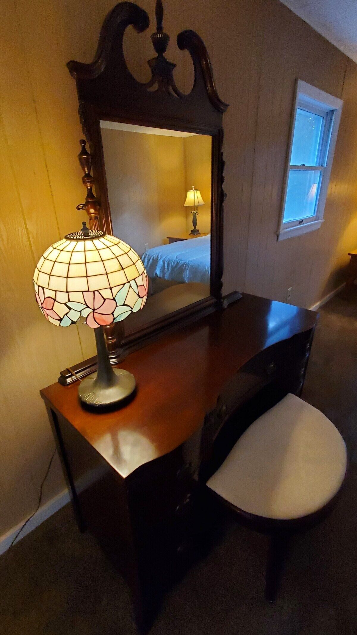 En byrå i trä med en spegel, en lampa med blommönster och en vit stol i ett rum med träväggar och ett fönster.