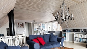 A modern living room with a high ceiling, a chandelier, a blue sofa with red cushions, a wooden coffee table, and a bookshelf filled with books.