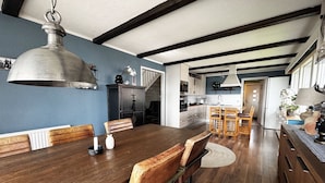 A dining area with a wooden table and chairs, a hanging light fixture, and a kitchen in the background.