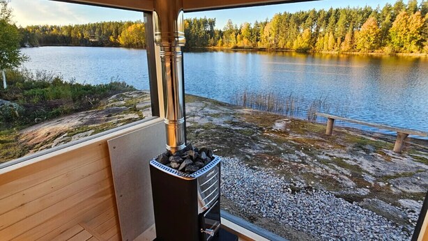 A wooden deck with a wood-burning stove, a view of a lake, and a forest in the background. - Newly built log cabin with sauna located on a lakeside plot outside Hultsfred | SE05086 (Vena)