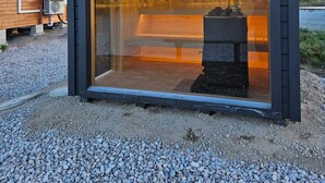 A modern glass-enclosed structure with a metal roof, situated on a gravel surface with a wooden building in the background. - Newly built log cabin with sauna located on a lakeside plot outside Hultsfred | SE05086 (Vena)