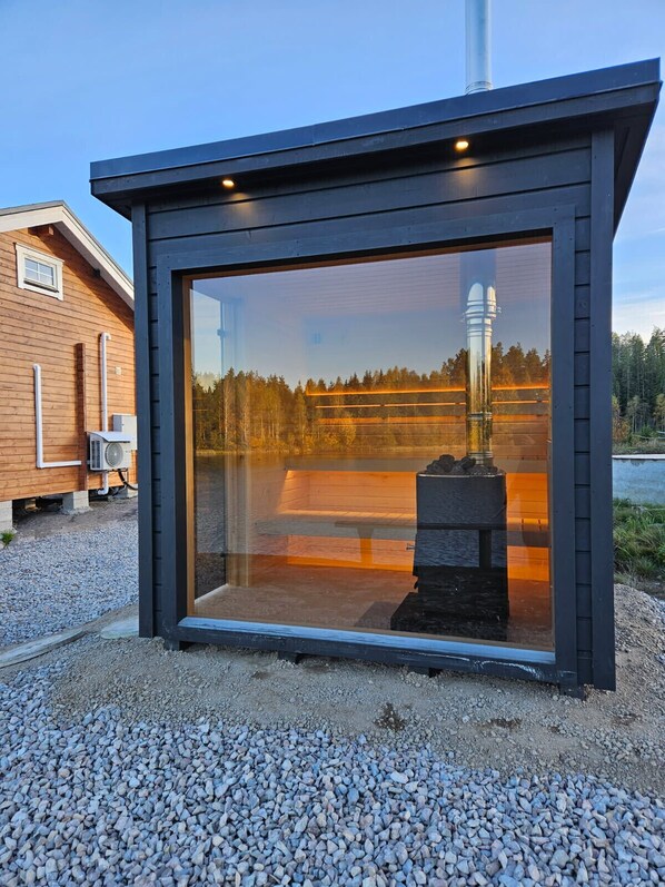 A modern glass-enclosed structure with a metal roof, situated on a gravel surface with a wooden building in the background.