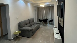 A modern living room with a grey sofa, a dining table with chairs, and a TV mounted on the wall.