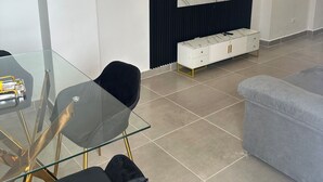 A modern living room with a glass dining table, black chairs, a grey sofa, a TV mounted on a black panel, and a white cabinet with gold legs.