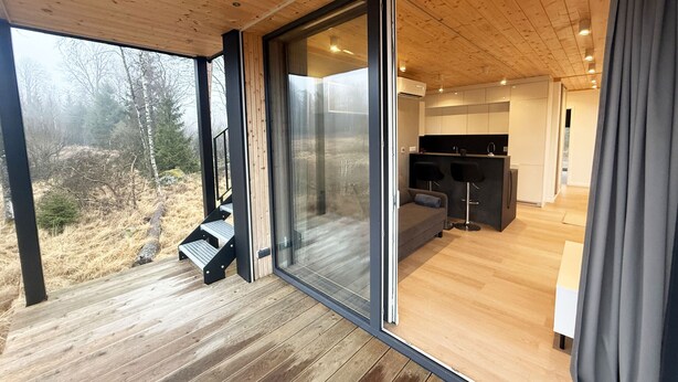 A modern interior with a wooden deck, a glass sliding door leading to a living area with a sofa and bar stools, and a view of the outdoors. - Holiday home in the middle of nature in Knäred |SE02014 (Knäred)
