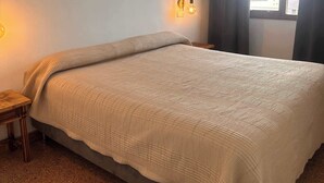 A neatly made bed with a quilted cover, a bedside table with a lamp, and a window with curtains.