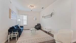 Una sala de estar moderna con un sofá azul, un sillón gris, un reposapiés blanco, una mesa de centro de vidrio y una alfombra estampada.