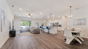 Un salón espacioso con un sofá en forma de L, un comedor con mesa y sillas blancas, y una cocina con armarios blancos y electrodomésticos modernos.