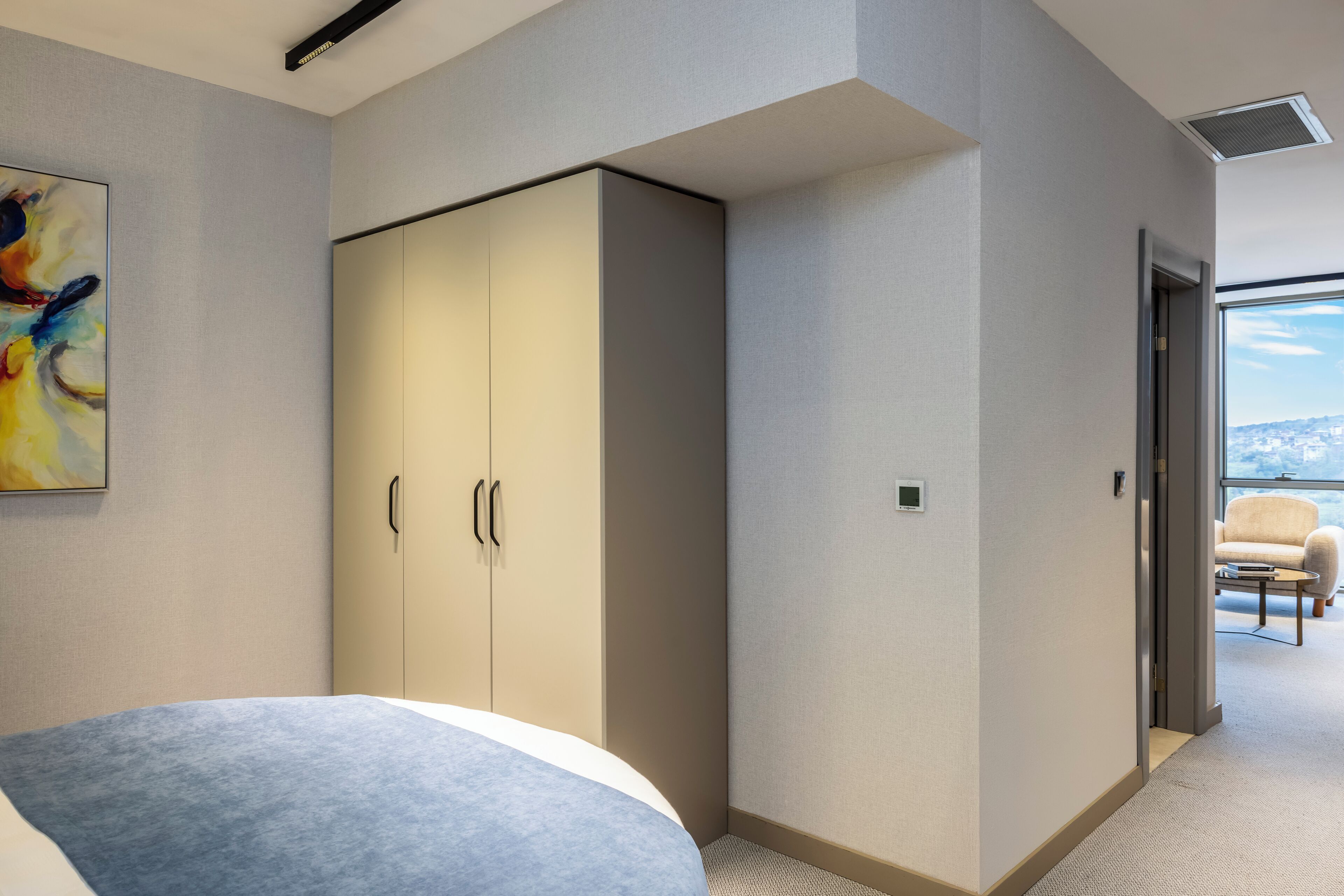 A modern bedroom with a large bed, a wardrobe, and a view of the city through a large window.