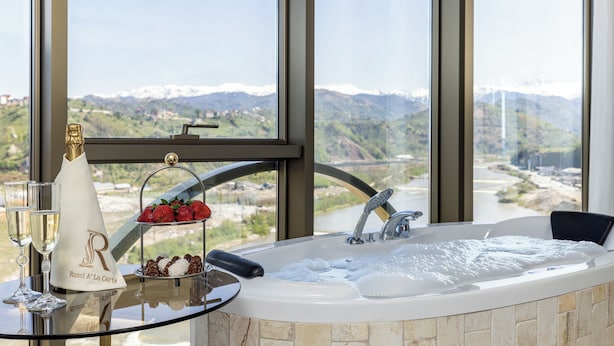 A luxurious bathroom with a large bathtub, a glass table with champagne and strawberries, and a view of mountains and a river. - Rossi Luxury Hotel (Ardesen)