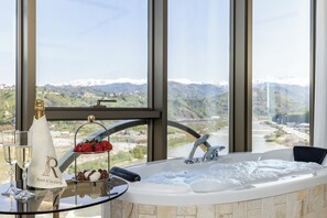 Une salle de bain luxueuse avec une grande baignoire, une table en verre sur laquelle sont disposés du champagne et des fraises, et une vue sur les montagnes et une rivière.
