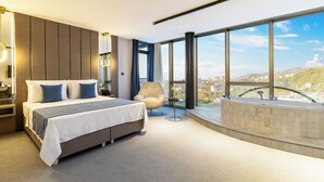 A modern hotel room with a large bed, a sitting area, and a city view through large windows. - Rossi Luxury Hotel (Ardesen)