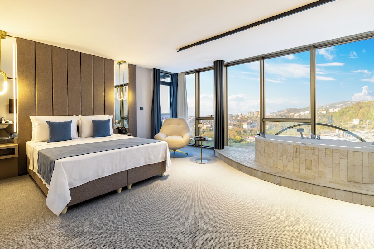 Ett modernt hotellrum med en stor säng, en sittgrupp och utsikt över staden genom stora fönster.
