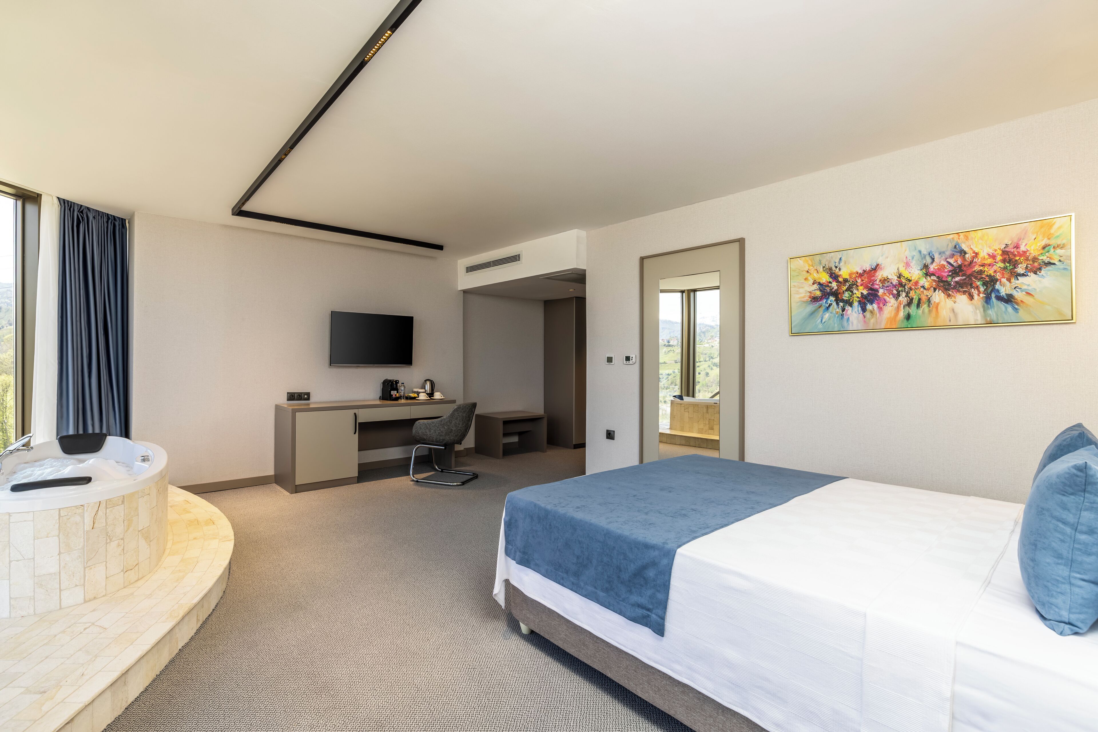 A modern hotel room with a large bed, a desk, a chair, a flat-screen TV, and a painting on the wall.