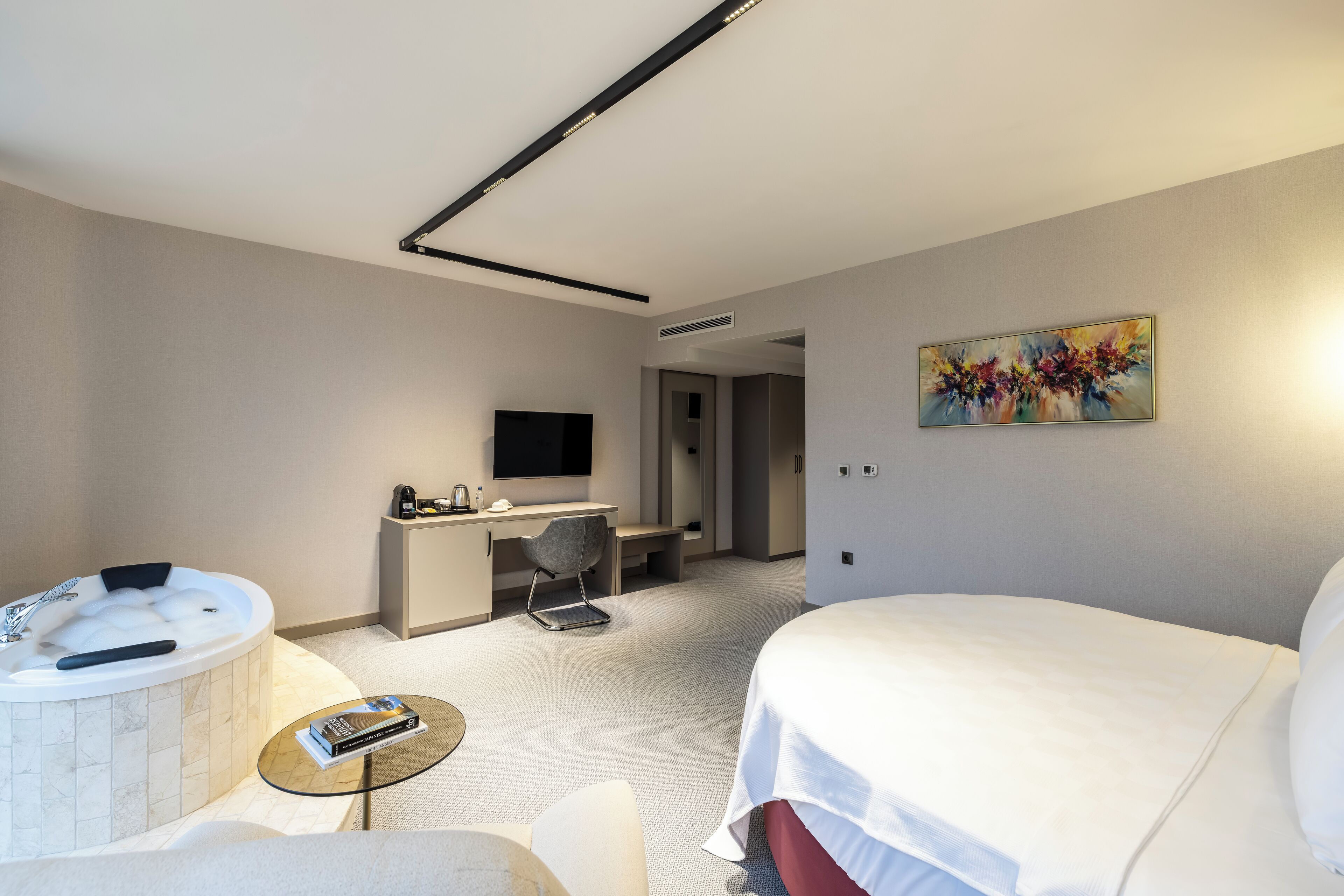 A modern hotel room with a large bed, a desk with a chair, a small round table with a book, a bathtub, and a painting on the wall.