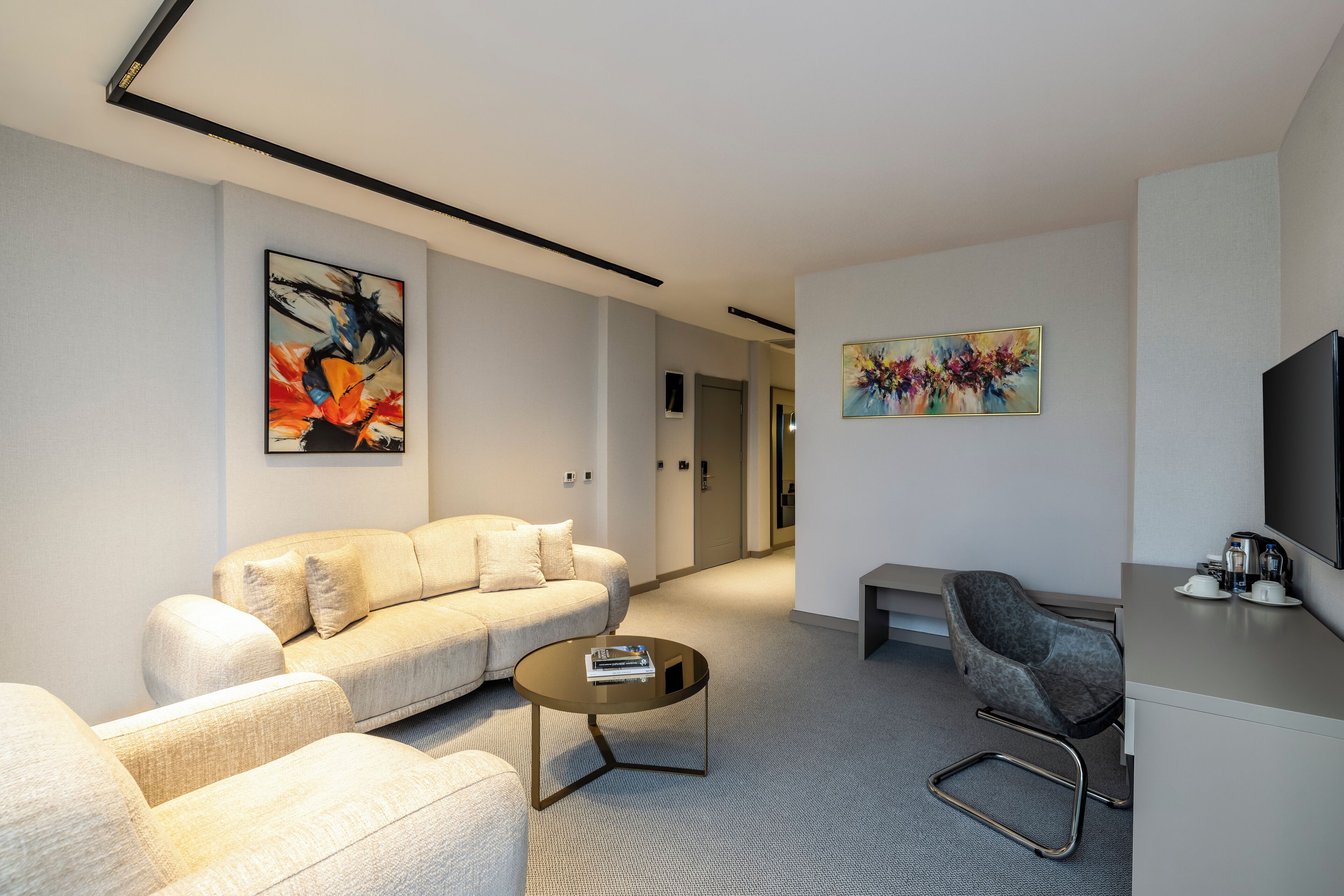 A modern living room with a sofa, coffee table, and a grey armchair. There are paintings on the walls and a flat-screen TV on a stand.