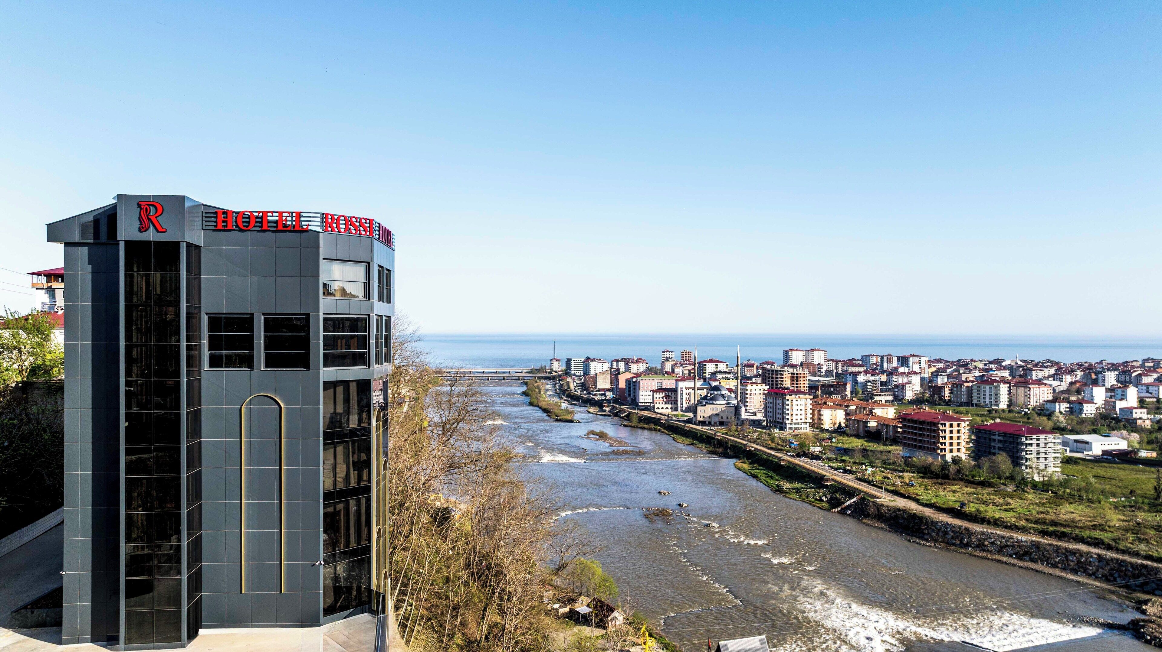 A modern hotel with a prominent 'HOTEL ROSS' sign, situated by a river with a view of a cityscape.