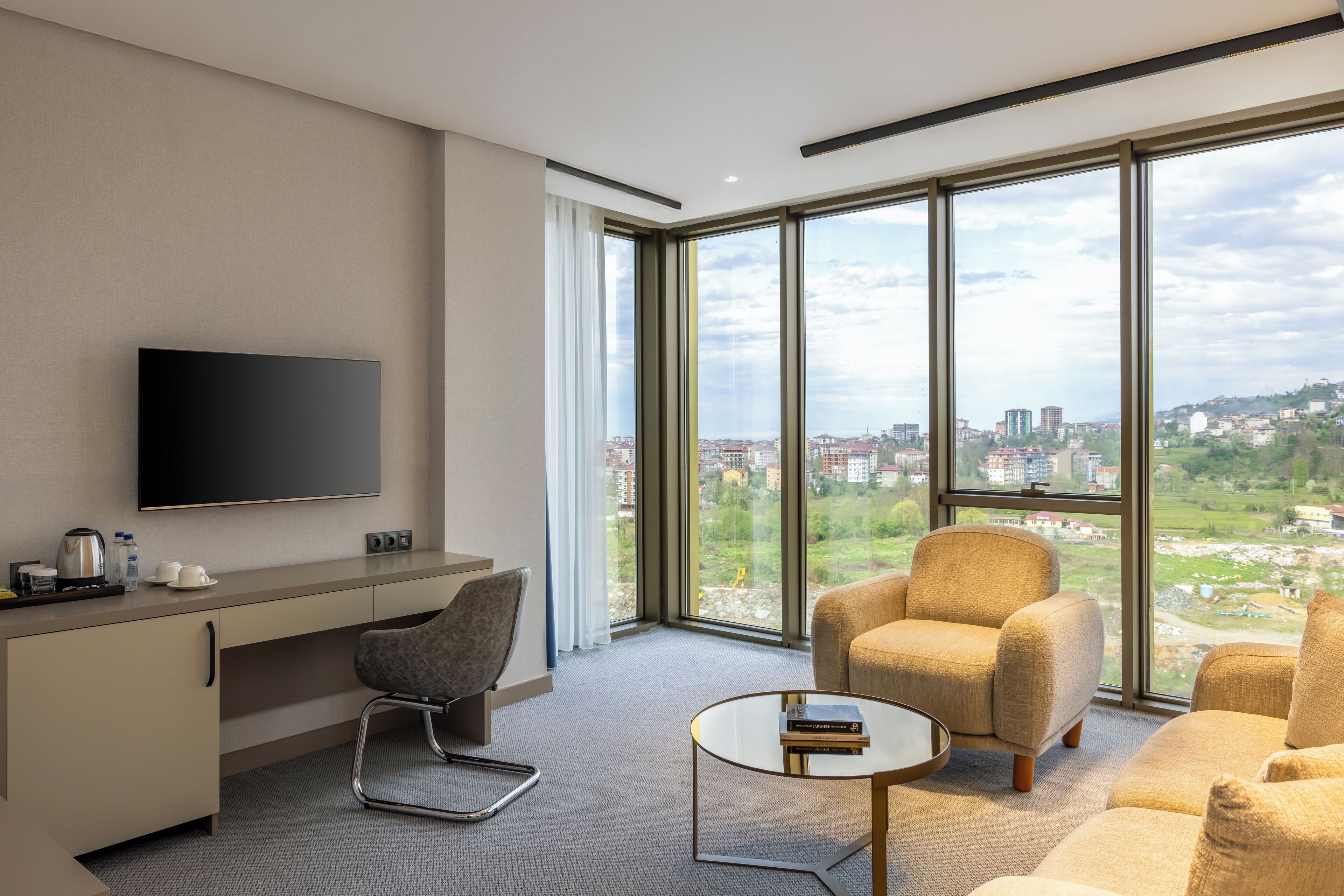 A modern hotel room with a large window offering a city view, a flat-screen TV, a desk with a chair, and a sofa.