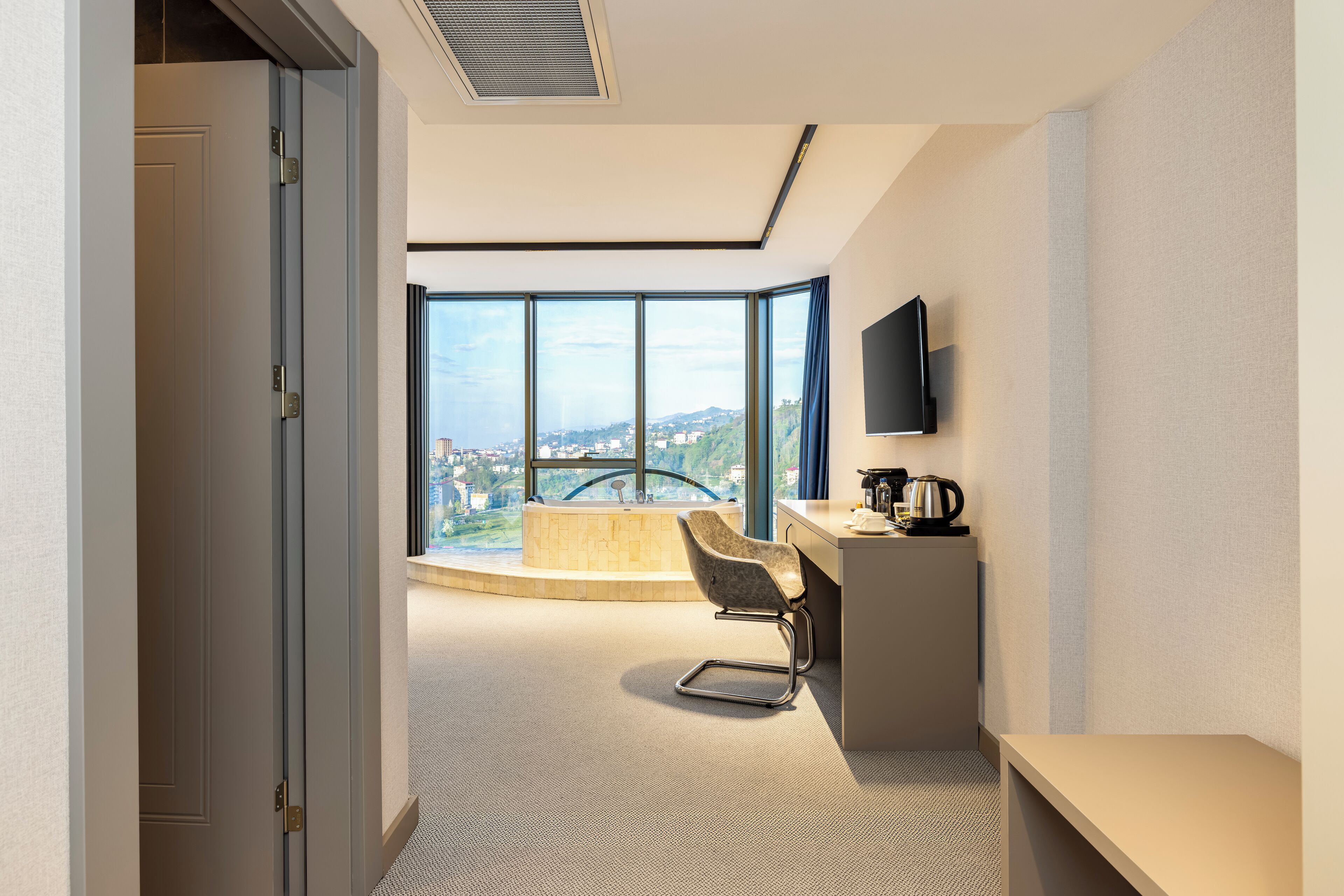 A modern hotel room with a large window offering a city view, a desk with a flat-screen TV, and a comfortable chair.