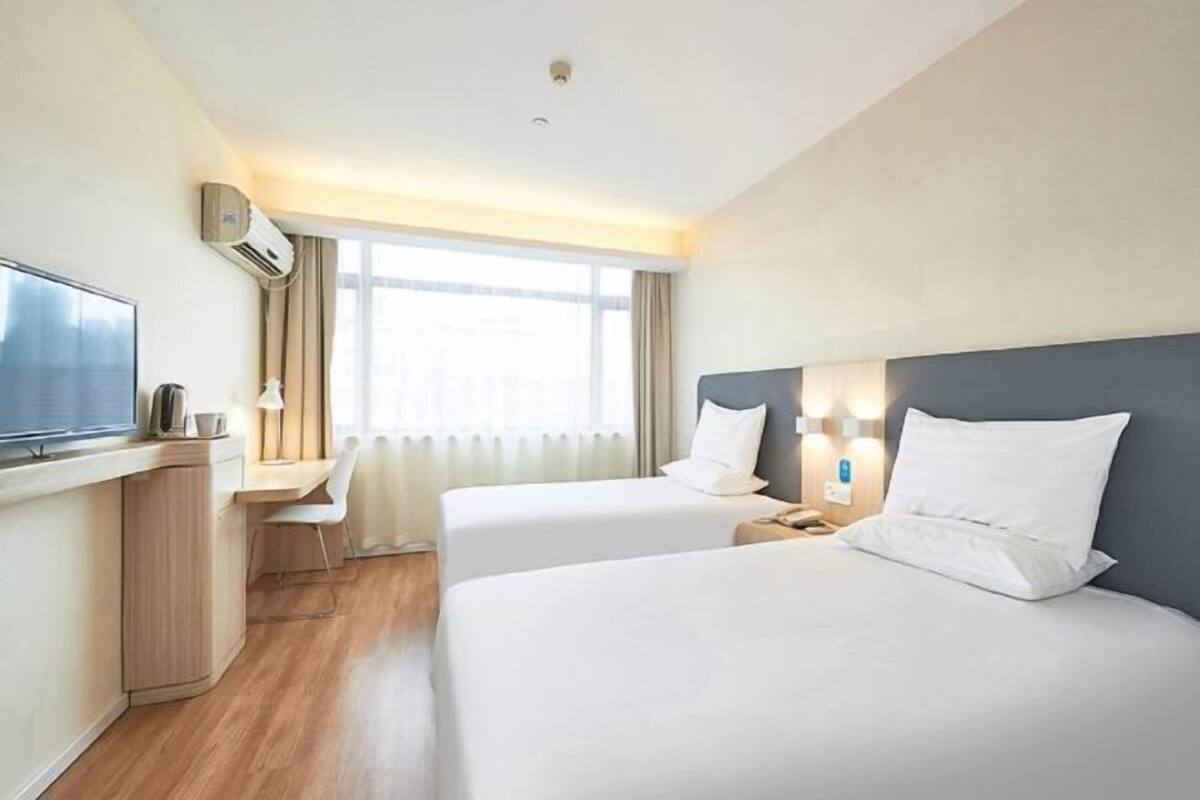 Quarto de hotel com duas camas, uma escrivaninha, televisão e janela com cortinas.