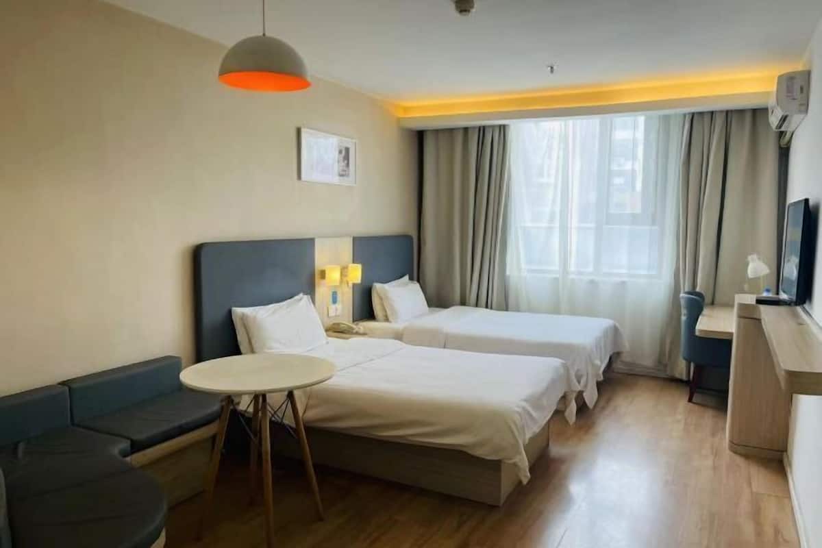 Quarto de hotel com duas camas, uma mesinha, um sofá, uma televisão e uma janela com cortinas.