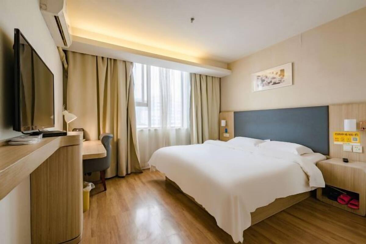 Quarto de hotel com cama, escrivaninha, cadeira, televisão e janela com cortinas.