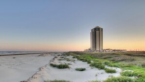 Paisatge costaner amb una platja sorrenca i dunes, un edifici alt al fons i un cel serè.