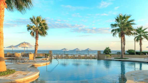 Zona de piscina amb gandules i ombrel·les, palmeres, i vista al mar i a les muntanyes.