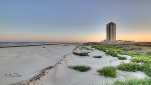 Paisaje costero con playa arenosa y dunas, un edificio alto al fondo, y cielo despejado.