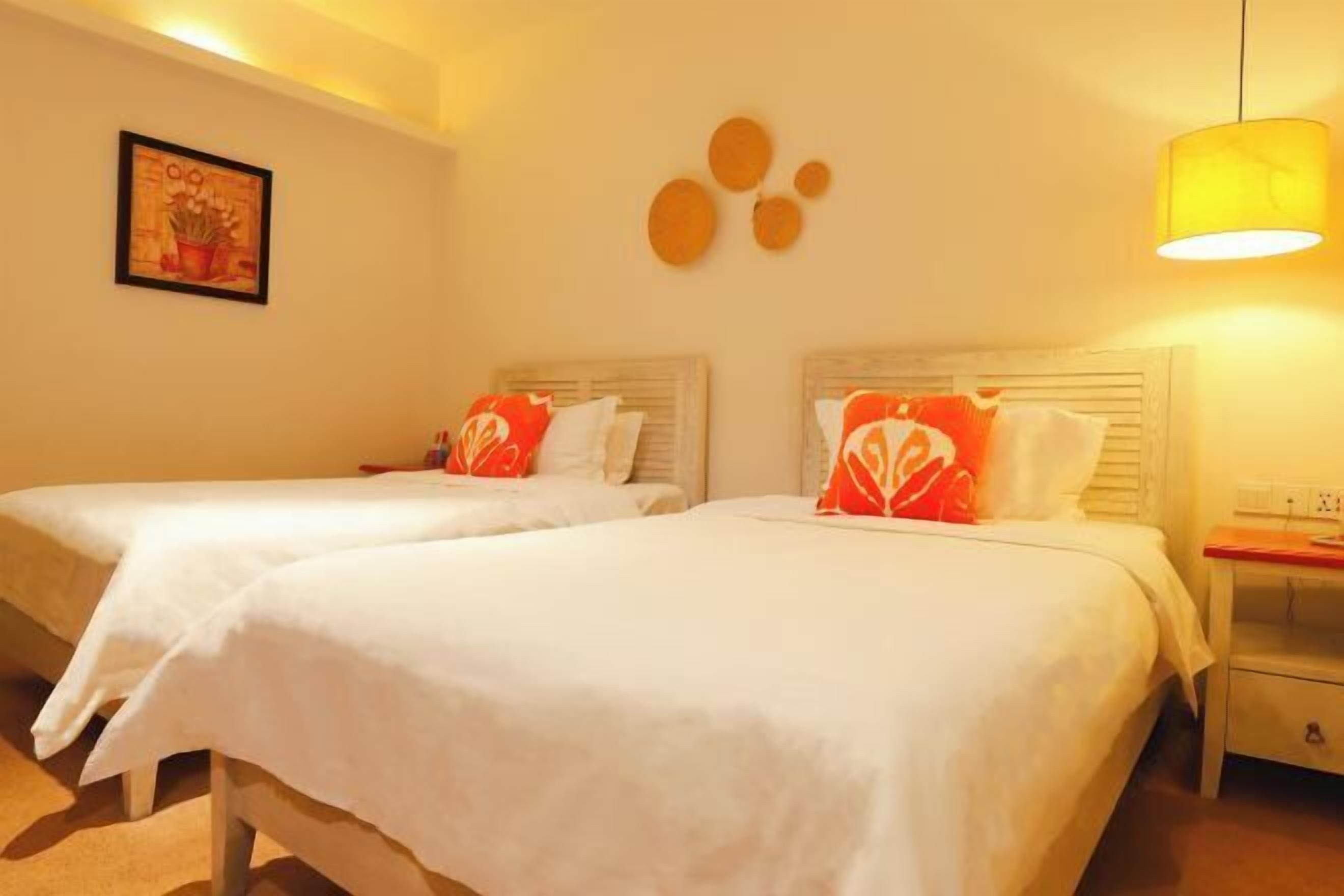A hotel room with two beds, each with white bedding and red decorative pillows. There is a small bedside table with a lamp on the right side.