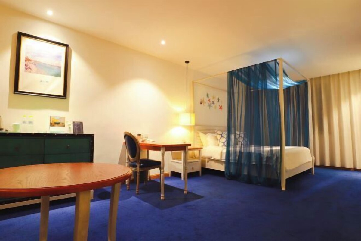 Habitación de hotel con cama, escritorio, silla, mesita de noche, lámpara, armario y un cuadro en la pared.