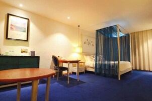 Habitación de hotel con cama, escritorio, silla, mesita de noche, lámpara, armario y un cuadro en la pared.