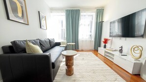 Een moderne woonkamer met een zwarte leren bank, een ronde houten salontafel, een witte console met een flatscreen televisie en een groot raam met groene gordijnen.