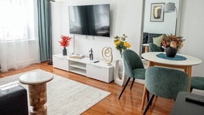 Een moderne woonkamer met een flatscreen televisie aan de muur, een witte tv-meubel, een ronde salontafel en groene beklede stoelen.