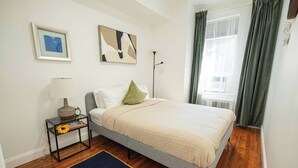 Een slaapkamer met een bed, een nachtkastje met een lamp, een lijst met een foto en een raam met gordijnen.