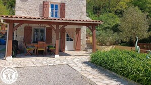 Ein Steinhaus mit Ziegeldach, eine überdachte Terrasse mit Tisch und Stühlen und eine Statue im Garten.