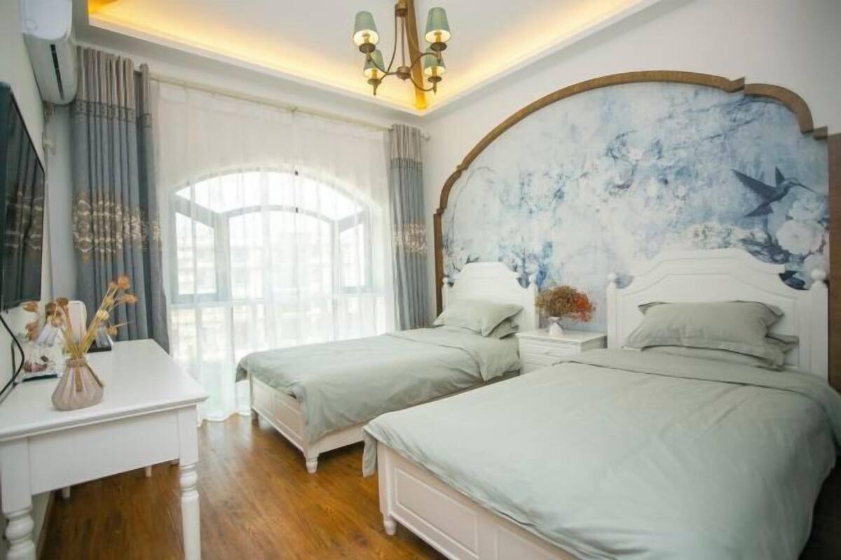 Quarto com duas camas, uma escrivaninha, um candelabro e uma janela grande.