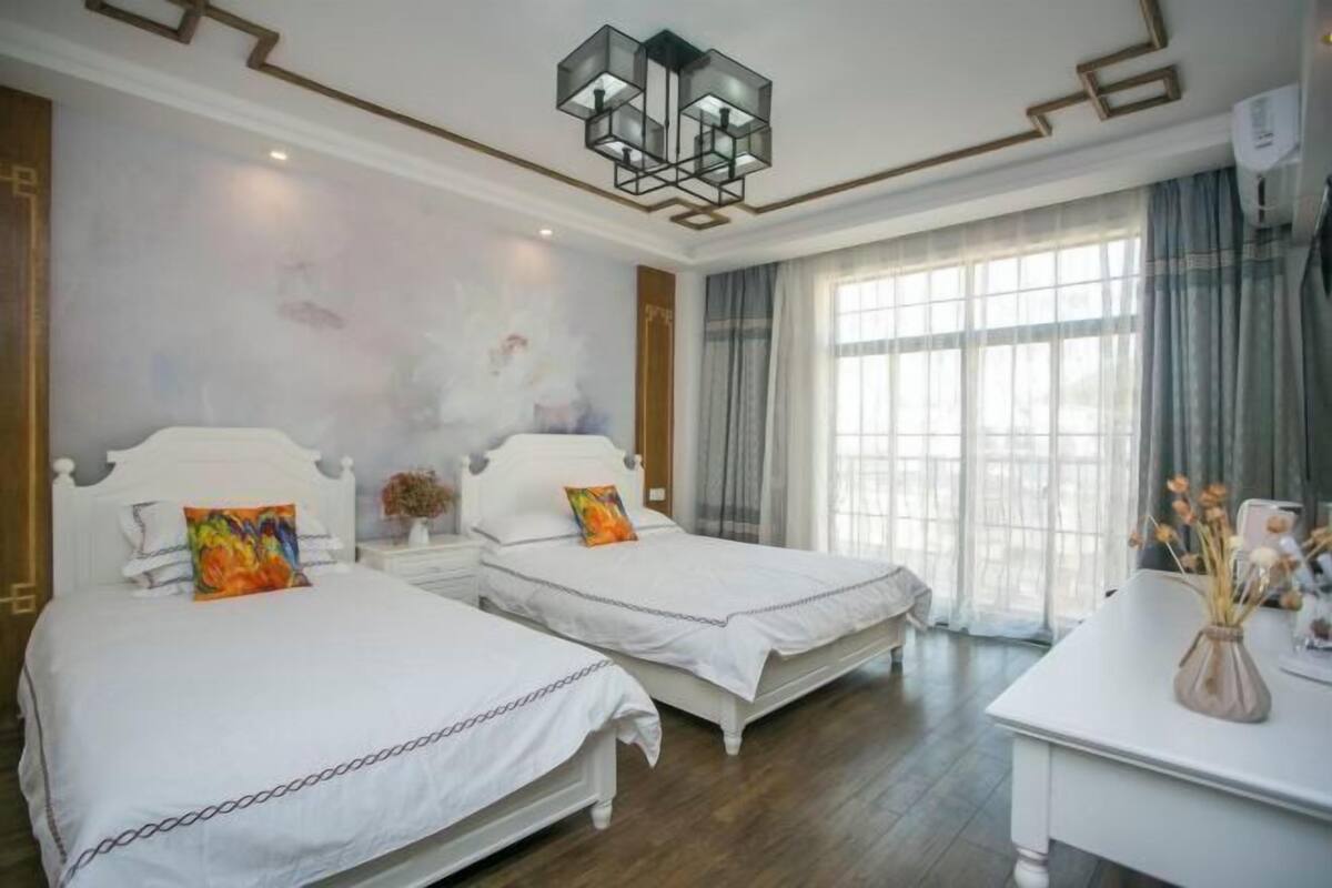 Quarto de hotel com duas camas, um candelabro, uma janela grande com cortinas e uma mesa branca com um vaso.
