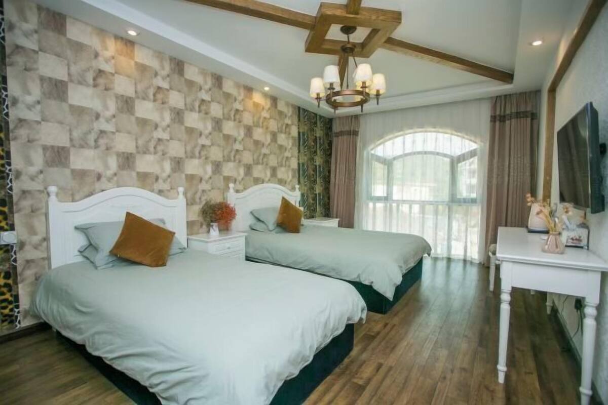 Quarto com duas camas, um candelabro, uma janela ampla e uma televisão.