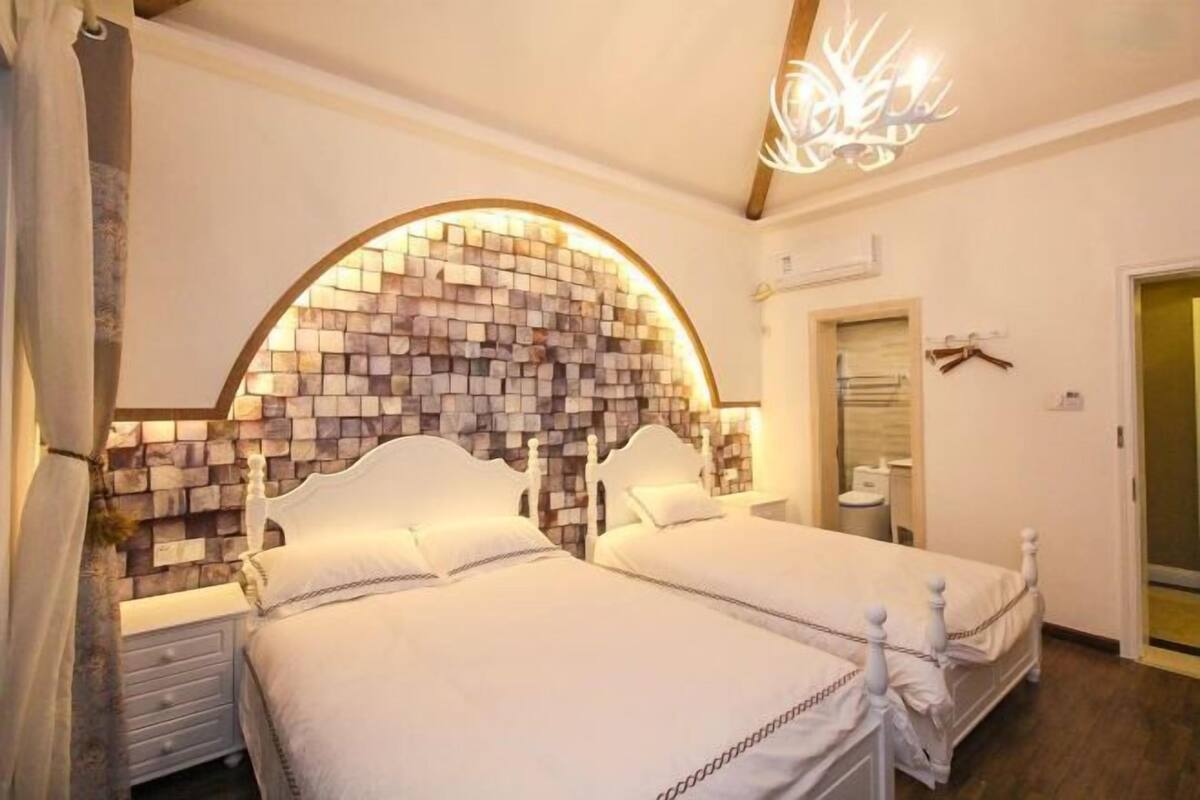 Quarto com duas camas, parede de pedra, lustre e banheiro pequeno.