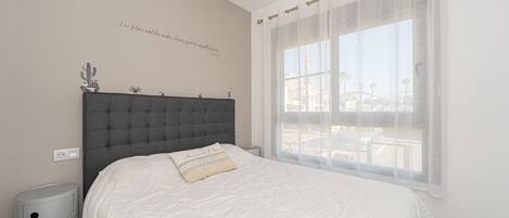 Un dormitorio con una cama grande, una mesita de noche, un enchufe en la pared y una ventana con cortinas ligeras.