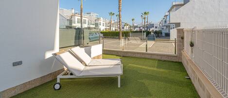 Un área de descanso al aire libre moderna, con una silla lounge blanca, césped artificial y vistas de los edificios circundantes.