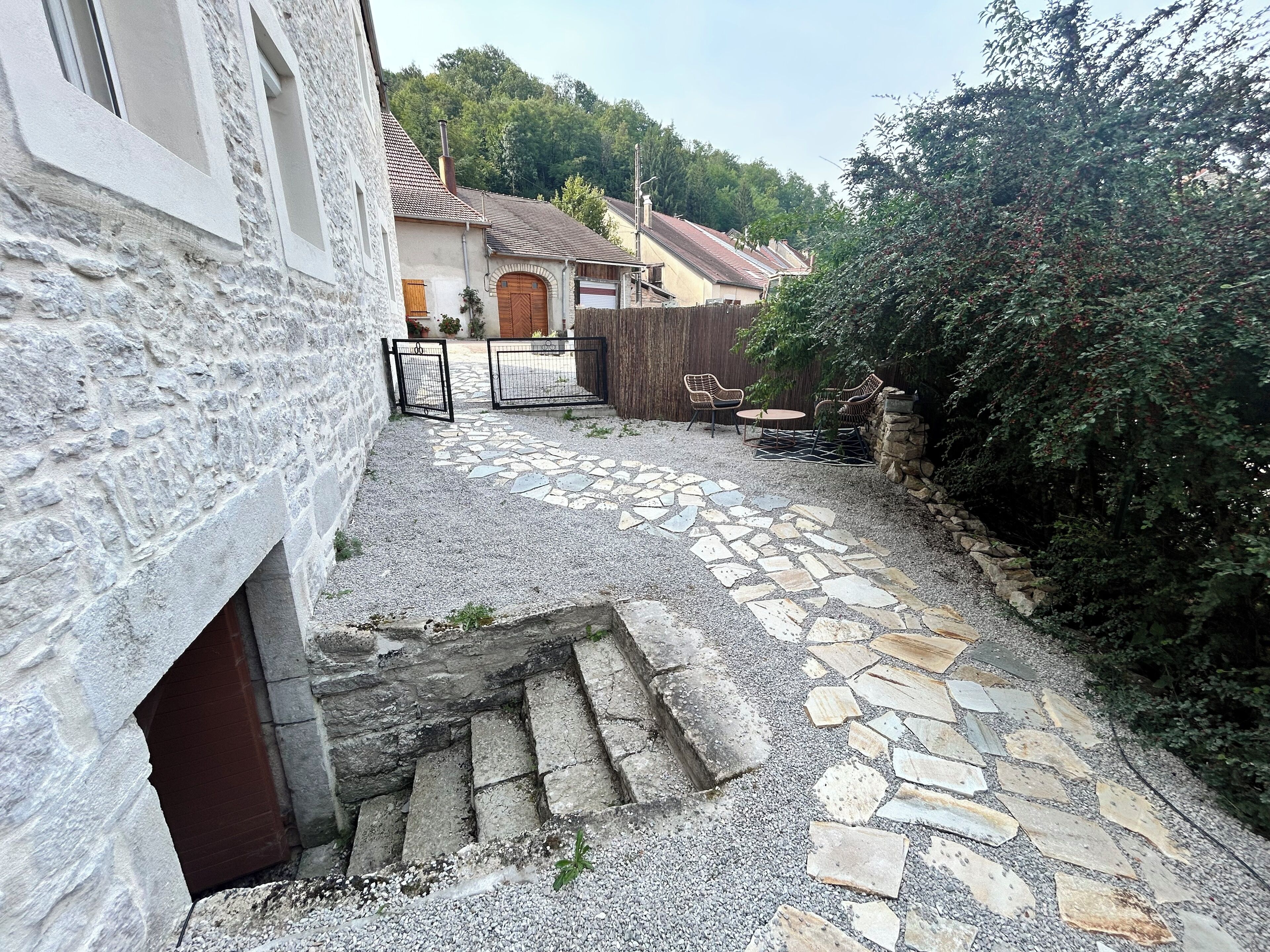 A stone pathway leading to a gated entrance with a wooden door, surrounded by a stone wall and greenery.