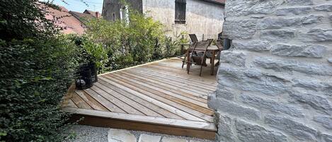 Une terrasse en bois, recouverte de carreaux de pierre, mène à un mur de pierre, le tout étant entouré de verdure et bordé par un bâtiment dont le toit est recouvert de tuiles rouges.