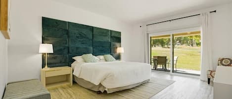 A bedroom with a large bed, a ceiling fan, a nightstand with a lamp, a bench, and a view of a grassy area through a sliding door.