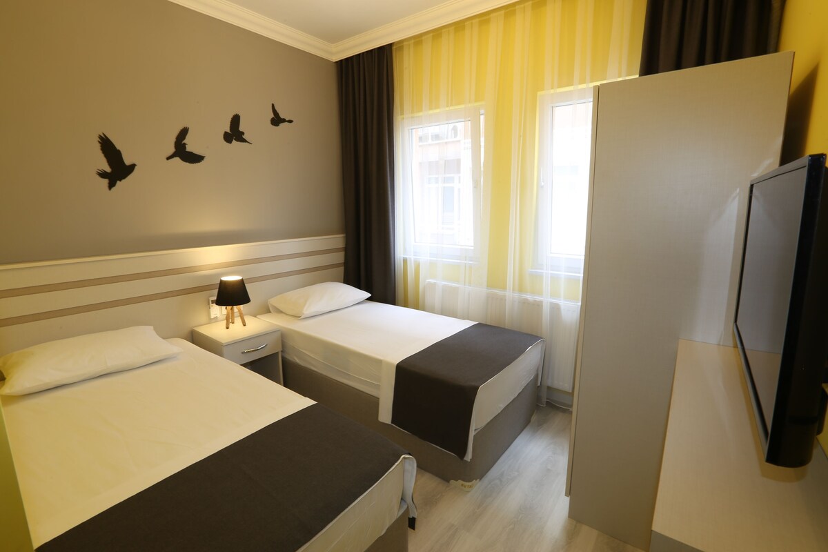 A hotel room with two beds, a nightstand with a lamp, a television, and a window with curtains.