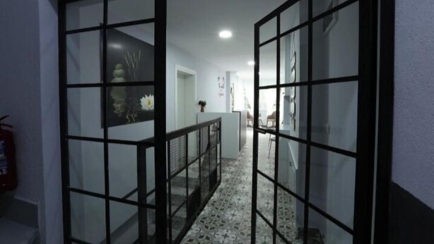 Un couloir doté de portes en verre et d’un sol à motifs, qui mène à une pièce comprenant un lit et une table de chevet.