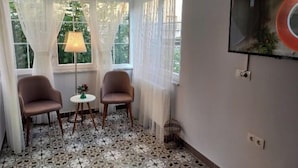 Une pièce comprenant deux fauteuils, une petite table et une lampe. On y trouve une grande fenêtre avec des rideaux, un téléviseur fixé au mur et un sol à motifs.