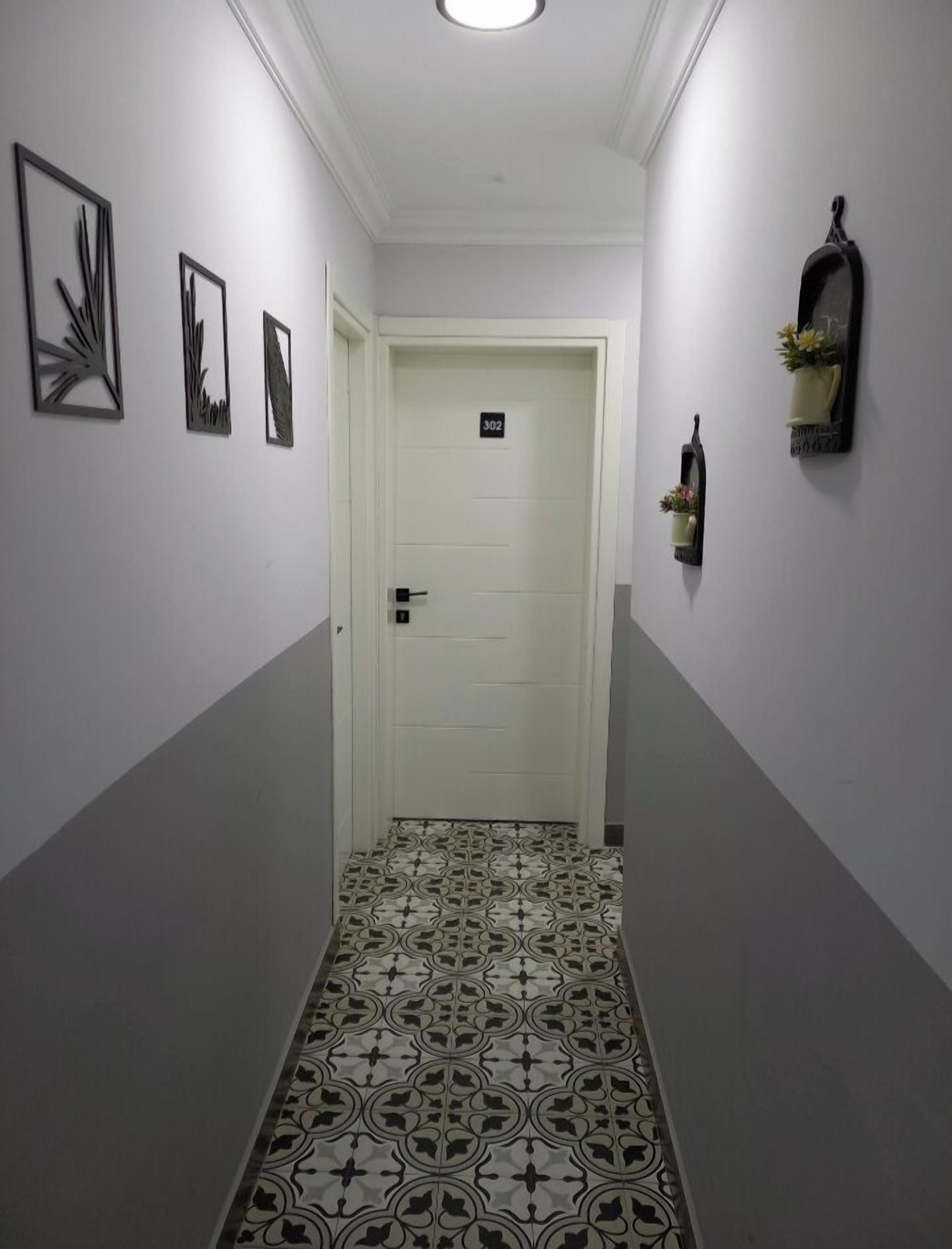 Un couloir étroit avec des carreaux de sol à motifs, une porte blanche portant un numéro, et des décorations murales, notamment des œuvres d’art encadrées et une petite étagère avec des fleurs.